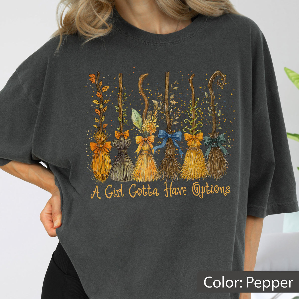 May include: Pepper-colored t-shirt featuring a whimsical design of six decorative brooms with colorful bows and floral accents. The text "A Girl Gotta Have Options" is printed below the brooms.