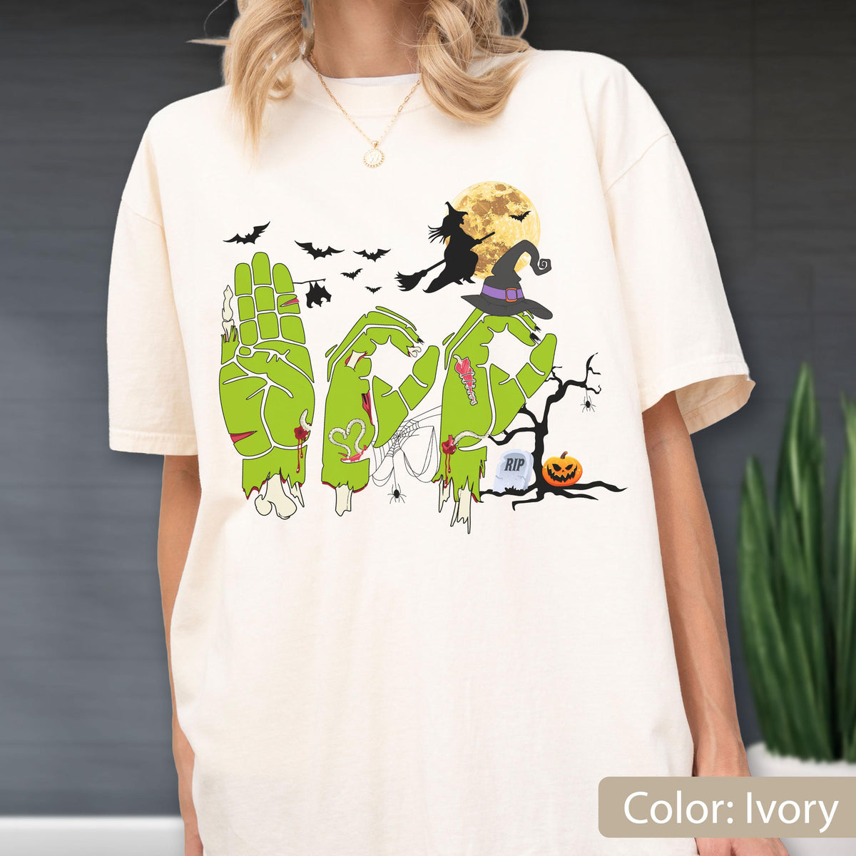 May include: An ivory t-shirt featuring a graphic of green zombie hands forming the word BOO. The design includes a silhouette of a witch on a broomstick, a full moon, bats, a spider web, a bare tree, a jack-o'-lantern, and a tombstone labeled RIP.