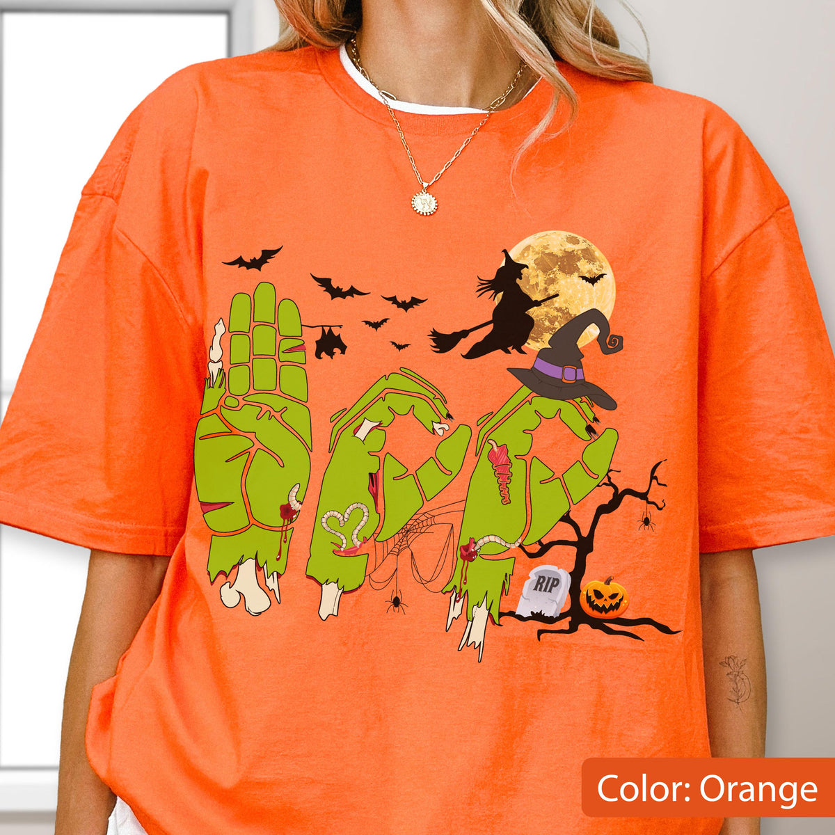May include: An orange t-shirt featuring a graphic of green zombie hands forming the word BOO. The design includes a full moon, a witch on a broomstick, bats, a spider web, a tombstone labeled RIP, and a jack-o'-lantern under a bare tree.