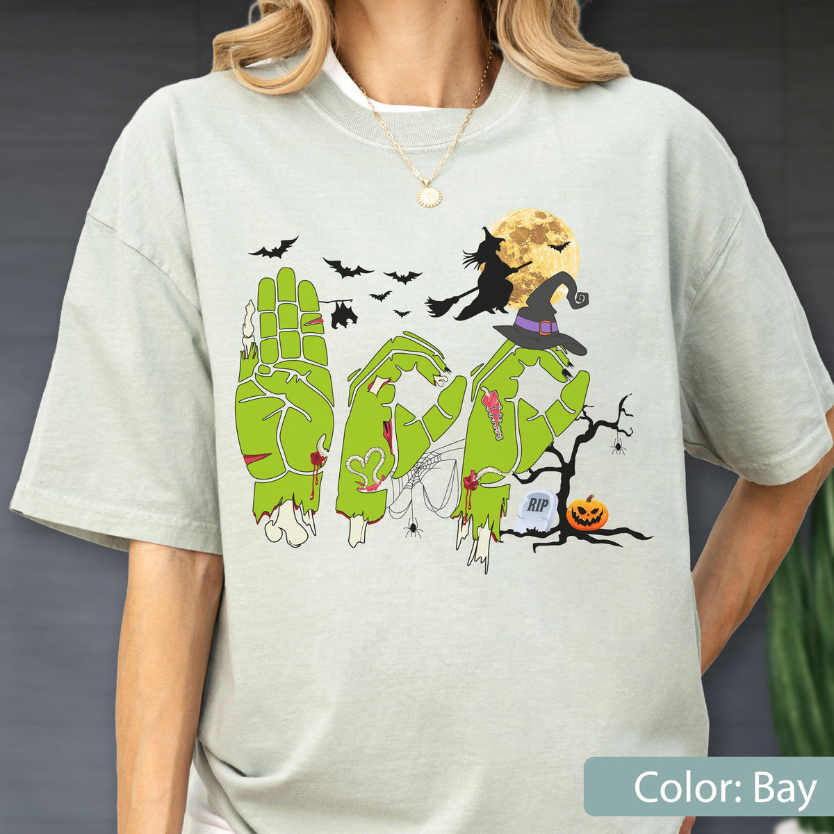 May include: A light green t-shirt featuring a graphic of three zombie hands forming the letters B-O-O in American Sign Language. The design includes a witch on a broomstick, a full moon, bats, a spider web, a tombstone with RIP, and a jack-o'-lantern.