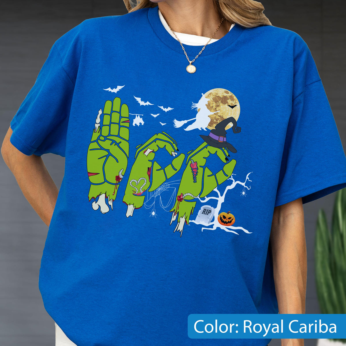 May include: A royal blue t-shirt featuring a graphic of green zombie hands forming the word BOO. The design includes a full moon, a witch on a broomstick, bats, a tombstone with RIP text, a jack-o'-lantern, and a bare tree against a dark background.