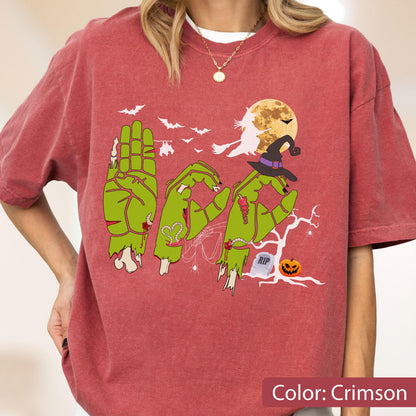 May include: A crimson short-sleeve t-shirt featuring a graphic of green zombie hands forming the word BOO. The design includes a witch on a broomstick, a full moon, bats, a witch hat, a tombstone with RIP text, a pumpkin, and a bare tree.