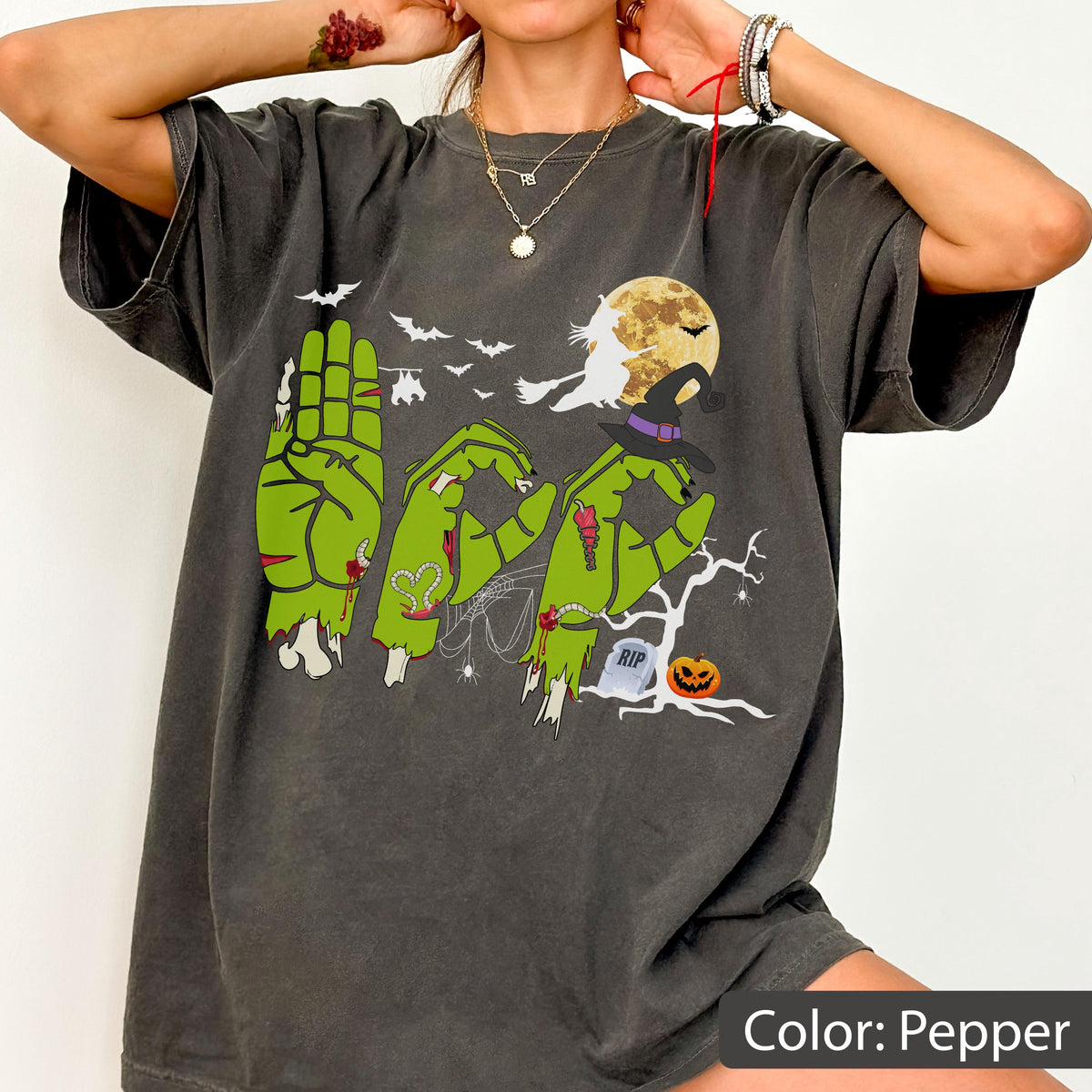 May include: A pepper-colored t-shirt featuring a graphic of green zombie hands forming the word BOO in sign language. The design includes a yellow full moon, a witch on a broomstick, bats, a bare tree, a tombstone marked RIP, and a jack-o'-lantern.
