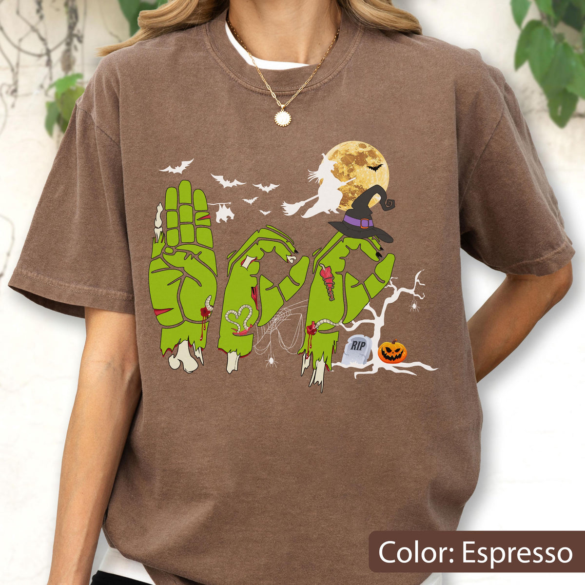 May include: An espresso brown t-shirt featuring a graphic of three green zombie hands forming the word BOO in American Sign Language. The design includes a yellow full moon, a witch on a broomstick, bats, a spider web, a tombstone, and a pumpkin.