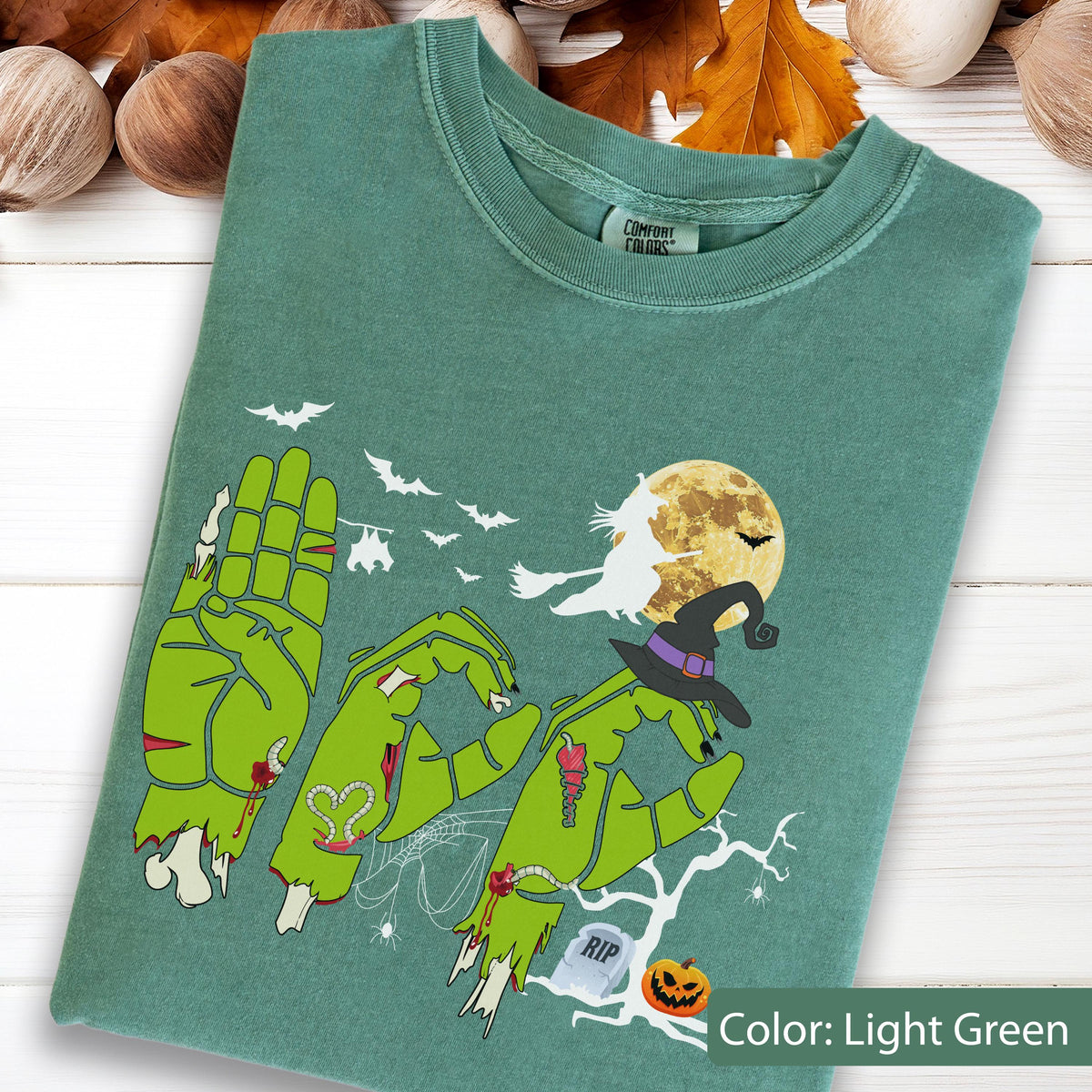 May include: A light green t-shirt featuring a graphic of three zombie hands forming the word BOO. The design includes a witch on a broomstick, a full moon, bats, a spider web, a tombstone with RIP text, a jack-o'-lantern, and a witch's hat.
