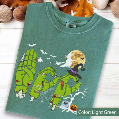 May include: A light green t-shirt featuring a graphic of three zombie hands forming the word BOO. The design includes a witch on a broomstick, a full moon, bats, a spider web, a tombstone with RIP text, a jack-o'-lantern, and a witch's hat.