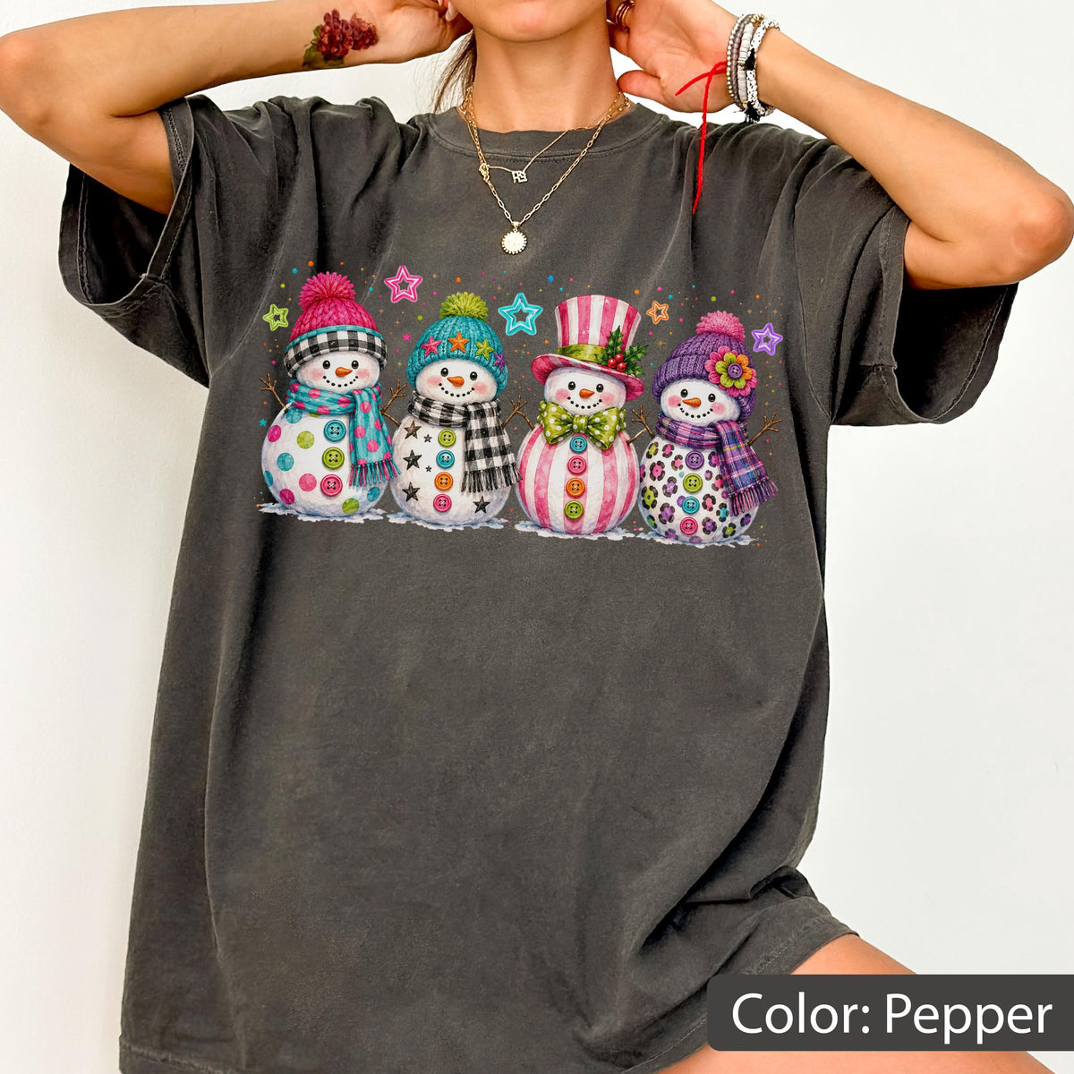 May include: A charcoal gray t-shirt featuring a graphic print of four snowmen in a row. Each snowman wears a colorful hat and scarf, decorated with buttons and patterns. The design includes small colorful stars scattered above the snowmen.