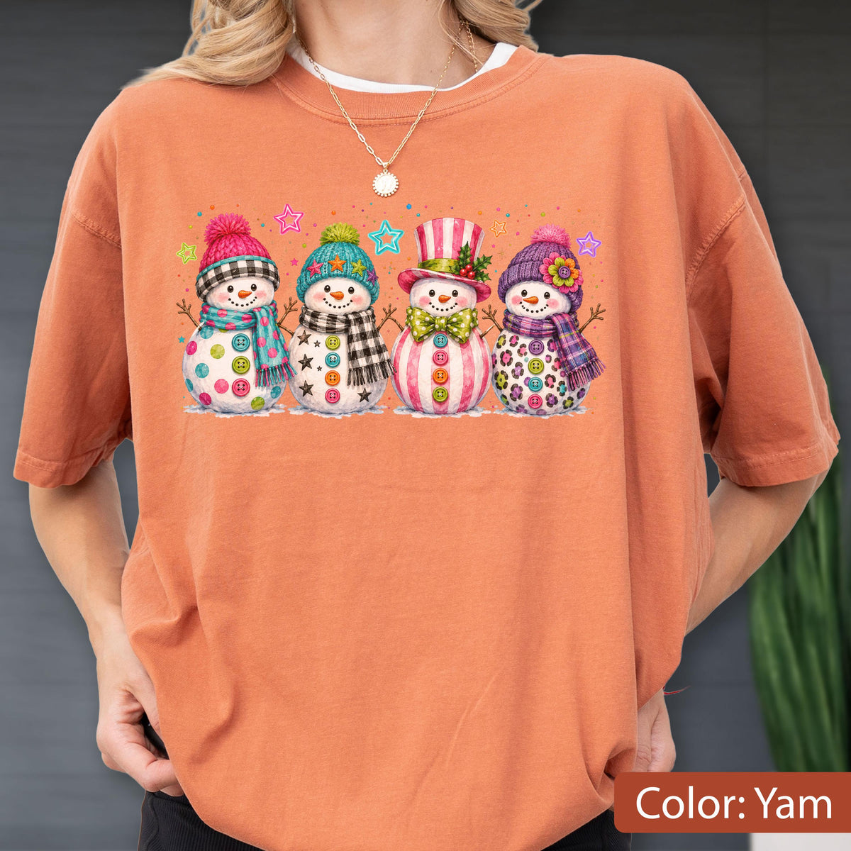 May include: A burnt orange short-sleeve t-shirt featuring a graphic print of four snowmen standing in a row. Each snowman wears a colorful hat and scarf, with various button designs on their bodies. Small colorful stars are scattered above them.