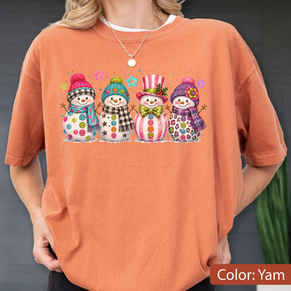 May include: A burnt orange short-sleeve t-shirt featuring a graphic print of four snowmen standing in a row. Each snowman wears a colorful hat and scarf, with various button designs on their bodies. Small colorful stars are scattered above them.