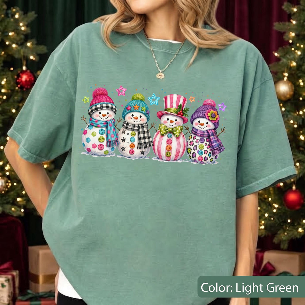 May include: A light green short-sleeve t-shirt featuring a graphic print of four snowmen wearing colorful hats and scarves, decorated with buttons and stars. The snowmen are arranged in a row, each with a unique winter-themed outfit and accessories.