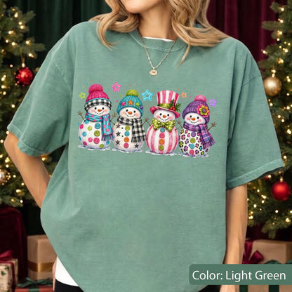 May include: A light green short-sleeve t-shirt featuring a graphic print of four snowmen wearing colorful hats and scarves, decorated with buttons and stars. The snowmen are arranged in a row, each with a unique winter-themed outfit and accessories.