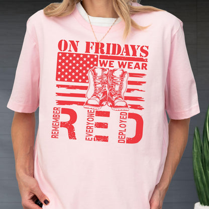 May include: A light pink t-shirt featuring a red graphic print of an American flag, a pair of military boots, and the text ON FRIDAYS WE WEAR RED REMEMBER EVERYONE DEPLOYED. The design is centered on the chest of the short-sleeved garment.