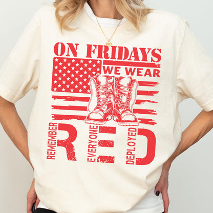 May include: A cream-colored short-sleeve t-shirt featuring a red graphic print. The design includes a stylized American flag, a pair of military boots, and the text: ON FRIDAYS WE WEAR RED, REMEMBER EVERYONE DEPLOYED.