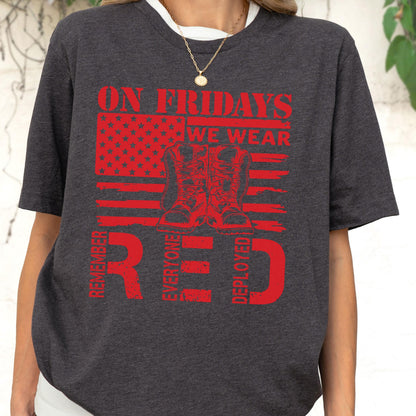 May include: A dark charcoal gray t-shirt featuring a red graphic print of an American flag with a pair of combat boots in the center. The text reads ON FRIDAYS WE WEAR RED and REMEMBER EVERYONE DEPLOYED in bold, distressed red lettering.
