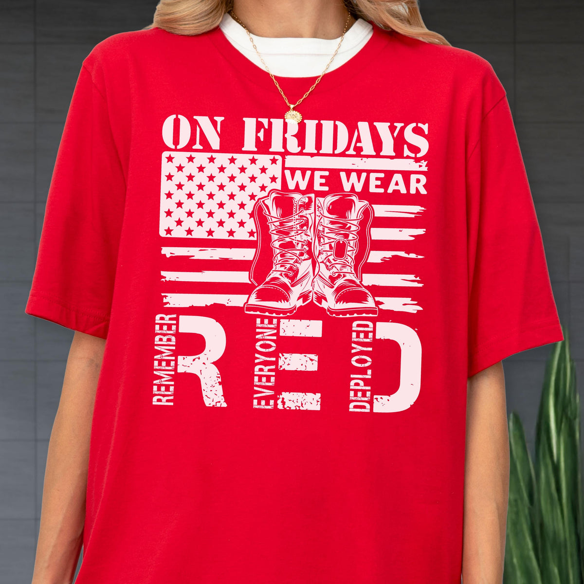 May include: A bright red t-shirt featuring a white graphic of the American flag, a pair of military boots, and the text ON FRIDAYS WE WEAR RED REMEMBER EVERYONE DEPLOYED. The shirt has a crew neckline and short sleeves, worn with a gold chain necklace.
