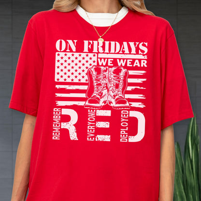 May include: A bright red t-shirt featuring a white graphic of the American flag, a pair of military boots, and the text ON FRIDAYS WE WEAR RED REMEMBER EVERYONE DEPLOYED. The shirt has a crew neckline and short sleeves, worn with a gold chain necklace.
