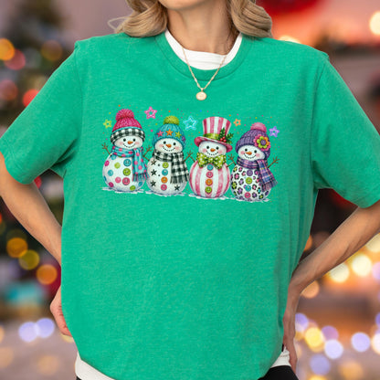 May include: A bright green short-sleeved t-shirt featuring a graphic print of four snowmen standing in a row. Each snowman wears a colorful winter hat and scarf, with decorative buttons on their bodies and small star shapes in the background.