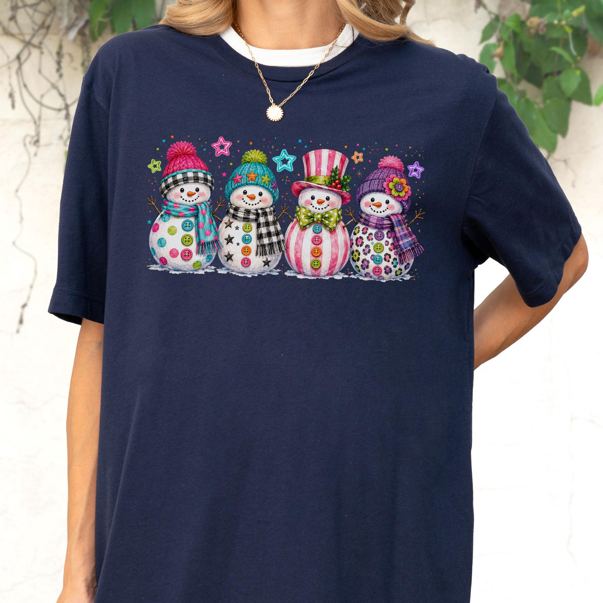 May include: A navy blue t-shirt featuring a graphic print of four snowmen standing in a row. Each snowman wears a colorful winter hat and scarf, with buttons on their bodies and small star shapes scattered in the background above them.