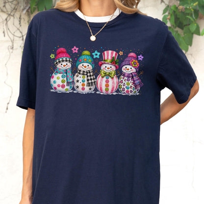 May include: A navy blue t-shirt featuring a graphic print of four snowmen standing in a row. Each snowman wears a colorful winter hat and scarf, with buttons on their bodies and small star shapes scattered in the background above them.