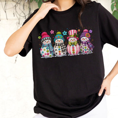May include: A black t-shirt featuring a graphic print of four snowmen standing in a row. Each snowman wears a colorful winter hat and scarf, with button details on their bodies. The design includes small star accents in various bright colors.