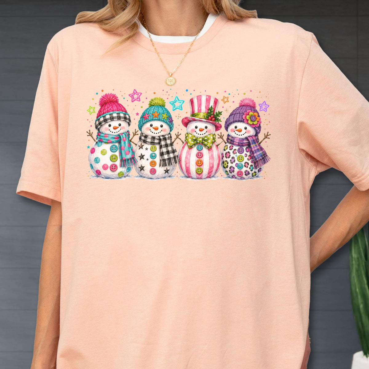 May include: A peach-colored t-shirt featuring a graphic print of four snowmen standing in a row. Each snowman wears a colorful winter hat and scarf, with various patterns like plaid, stripes, and floral accents, surrounded by small stars.