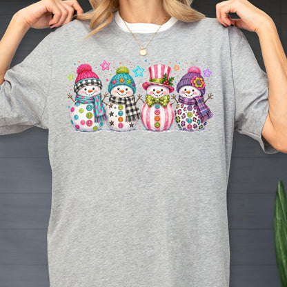 May include: A heather gray t-shirt featuring a graphic print of four snowmen standing in a row. Each snowman wears a colorful winter hat and scarf, with various patterns including plaid, stripes, and polka dots, accented by small colorful stars.