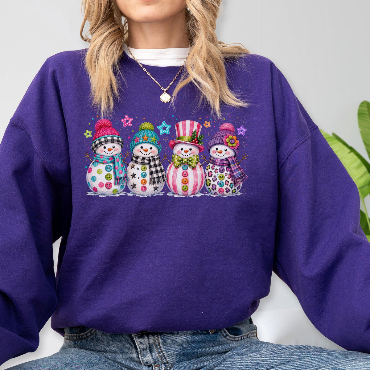 May include: A deep purple long-sleeve sweatshirt featuring a graphic print of four snowmen standing in a row. Each snowman wears a colorful hat and scarf with distinct patterns, including polka dots, stars, stripes, and leopard print designs.