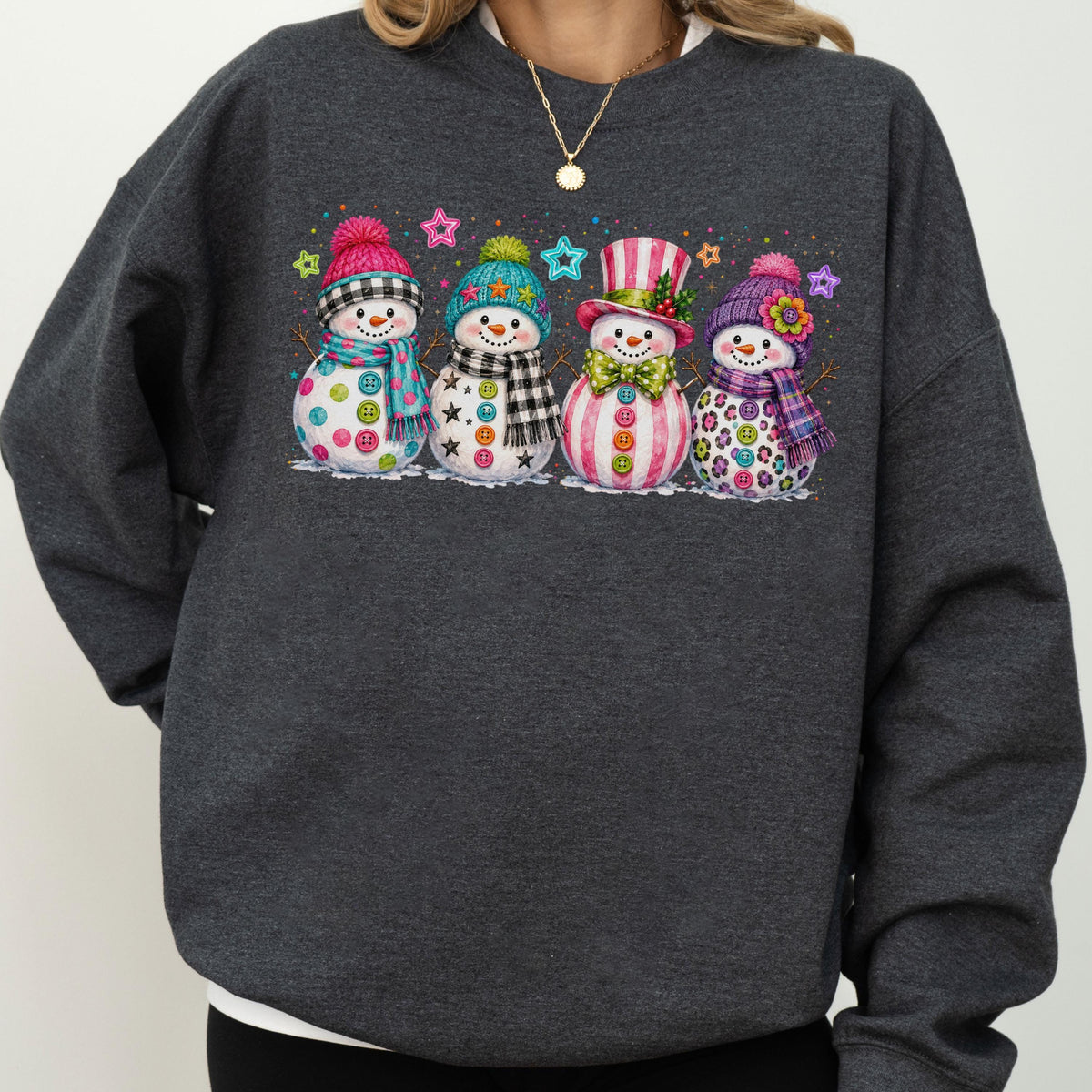 May include: A dark charcoal gray crewneck sweatshirt featuring a graphic print of four snowmen standing in a row. Each snowman wears a colorful hat and scarf, decorated with various buttons and small star accents in bright, festive colors.