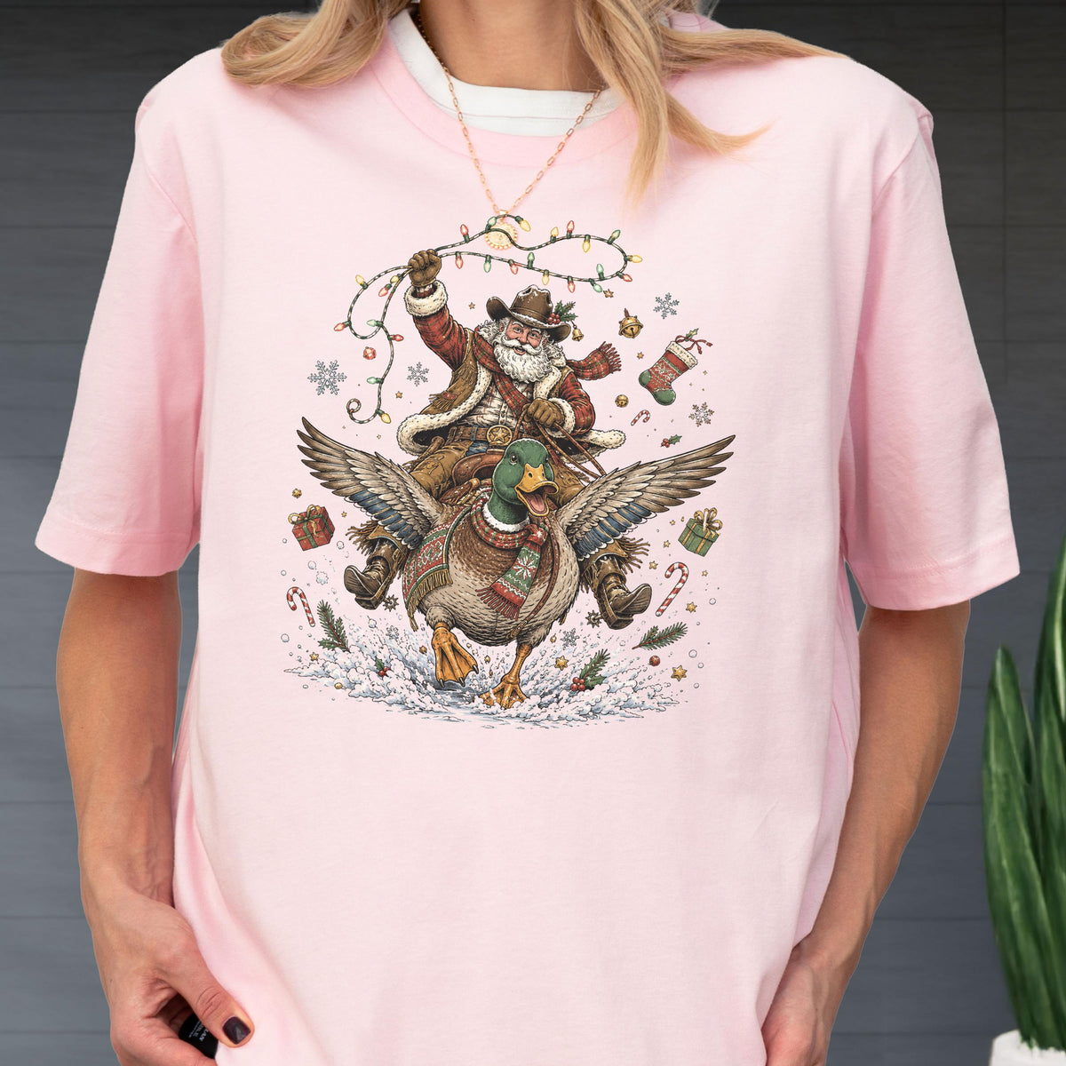 May include: A light pink t-shirt featuring a graphic of Santa Claus wearing a cowboy hat and vest while riding a flying mallard duck. The design includes Christmas lights, snowflakes, wrapped gifts, candy canes, and a stocking in the scene.