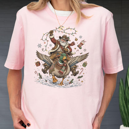 May include: A light pink t-shirt featuring a graphic of Santa Claus wearing a cowboy hat and vest while riding a flying mallard duck. The design includes Christmas lights, snowflakes, wrapped gifts, candy canes, and a stocking in the scene.