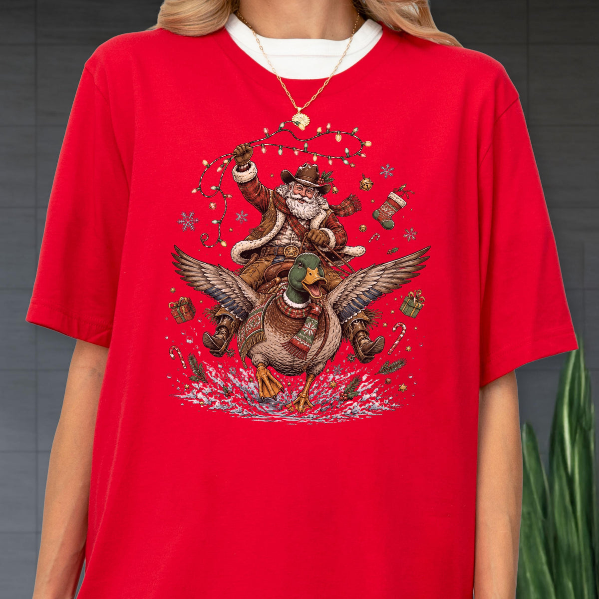 May include: A bright red t-shirt featuring a graphic illustration of Santa Claus riding a flying mallard duck. The design includes festive elements like string lights, snowflakes, gift boxes, and a stocking, all centered on the chest of the shirt.