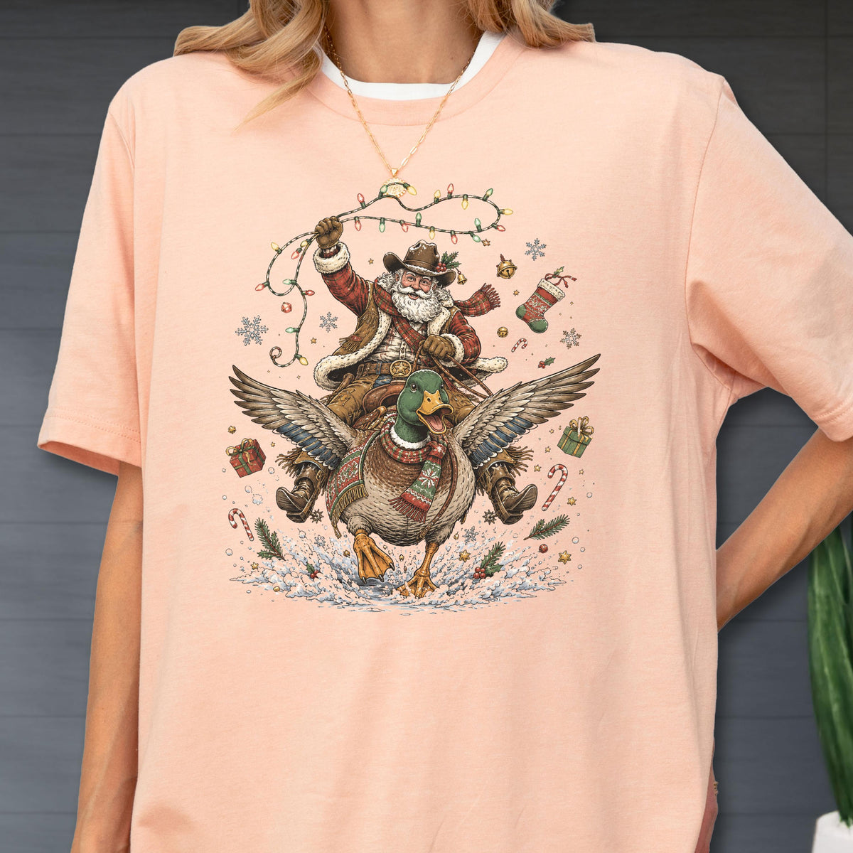 May include: A peach-colored t-shirt featuring a graphic print of Santa Claus wearing a cowboy hat and vest, riding a mallard duck. Santa holds a string of Christmas lights, surrounded by snowflakes, gift boxes, a stocking, and candy canes.