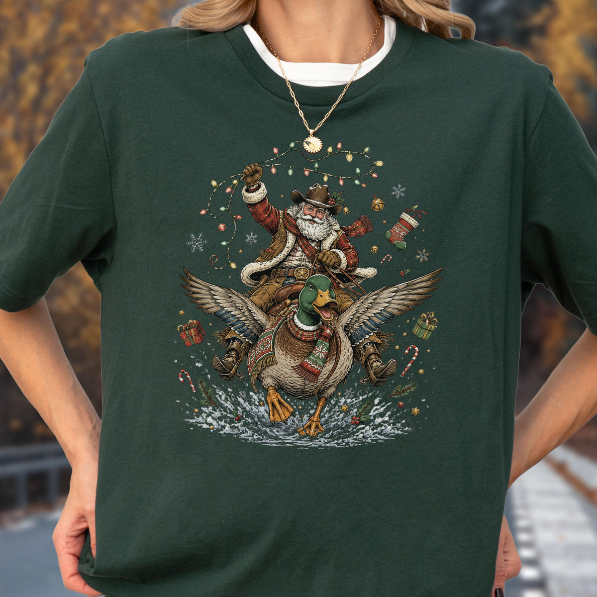 May include: A dark green short-sleeve t-shirt featuring a graphic print of Santa Claus wearing a cowboy hat and riding a flying mallard duck. The design includes Christmas lights, snowflakes, candy canes, and gift boxes surrounding the figures.