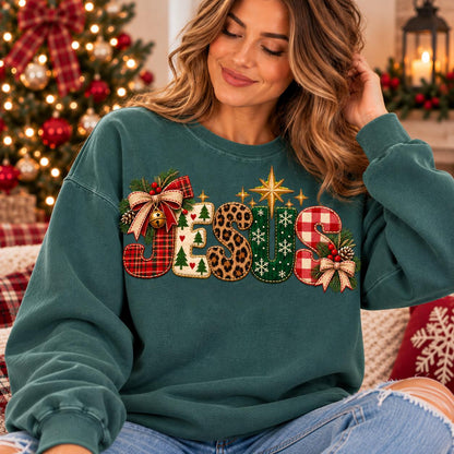 May include: A dark teal crewneck sweatshirt featuring the word JESUS in large, decorative letters. Each letter has a unique pattern including red plaid, white with green trees, leopard print, green with white snowflakes, and red gingham, with bows.