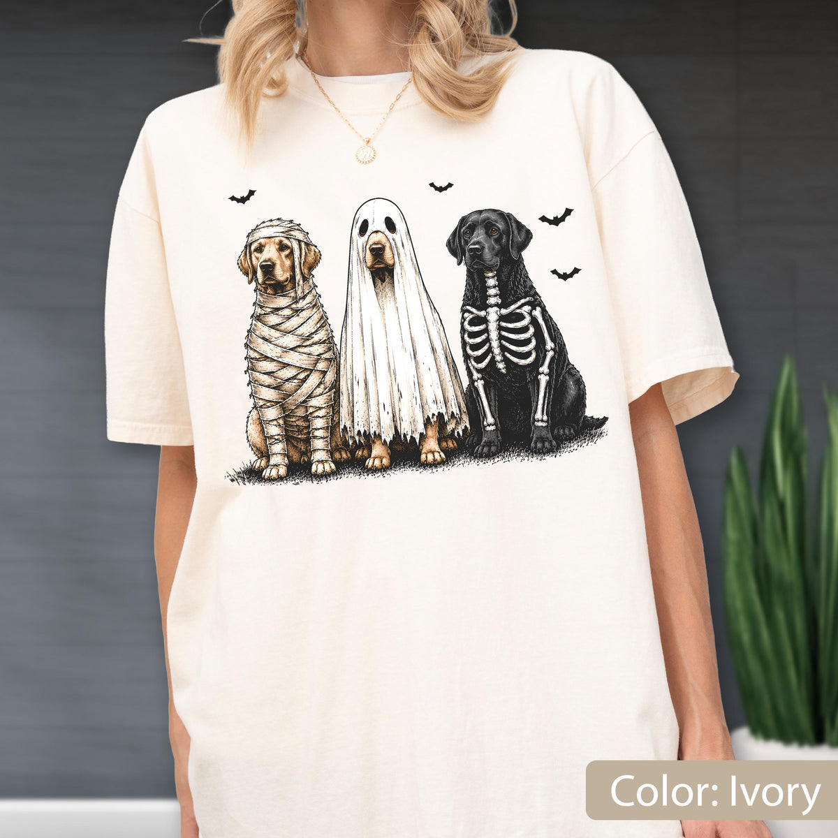May include: An ivory short-sleeve t-shirt featuring a graphic print of three dogs dressed for Halloween. One dog is wrapped like a mummy, one is wearing a white ghost sheet, and one has a skeleton ribcage design. Small black bats fly above them.