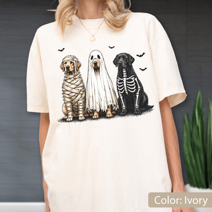 May include: An ivory short-sleeve t-shirt featuring a graphic print of three dogs dressed for Halloween. One dog is wrapped like a mummy, one is wearing a white ghost sheet, and one has a skeleton ribcage design. Small black bats fly above them.