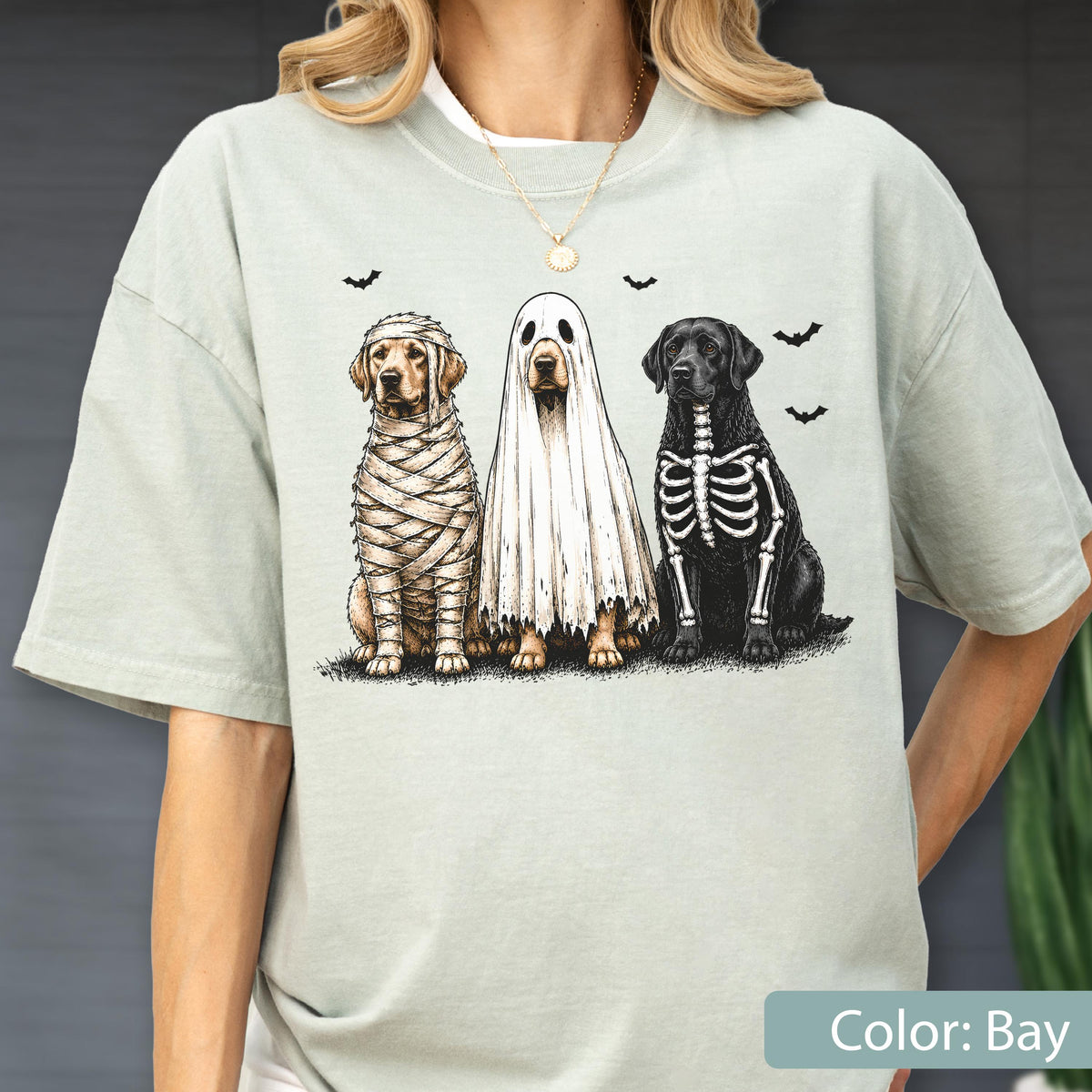 May include: A light sage green t-shirt featuring a graphic of three dogs in Halloween costumes. One dog is wrapped like a mummy, one is wearing a white ghost sheet, and one has a skeleton print on its black fur, with small black bats above.