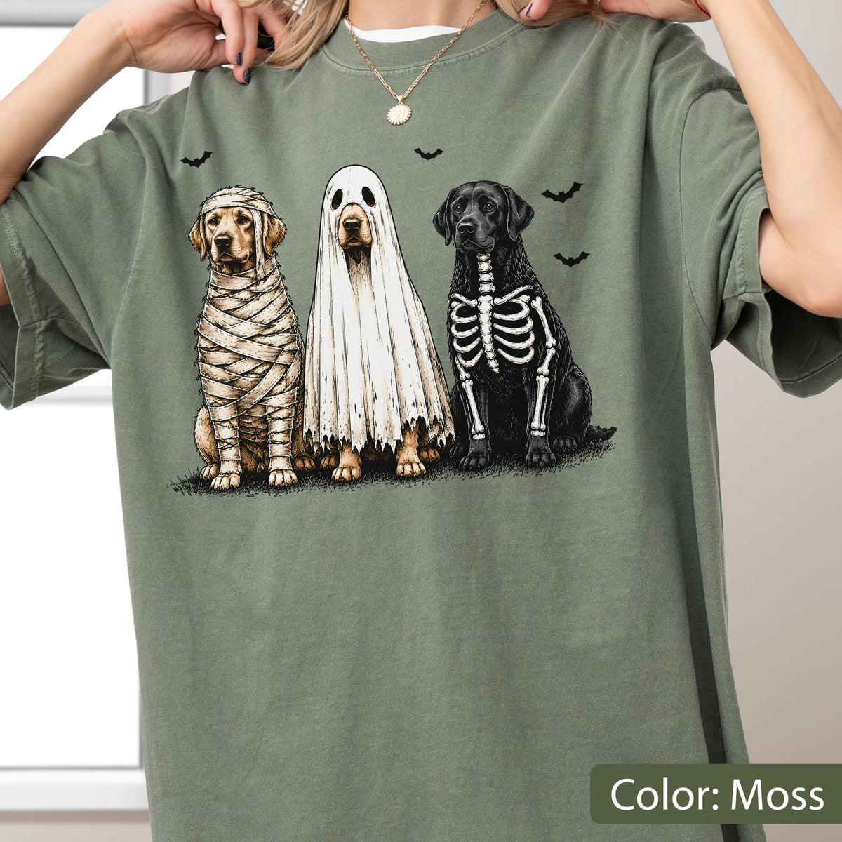 May include: A moss green t-shirt featuring a graphic print of three dogs in Halloween costumes. One dog is wrapped like a mummy, one is wearing a white ghost sheet, and one has a skeleton print on its body. Small black bats are printed above.