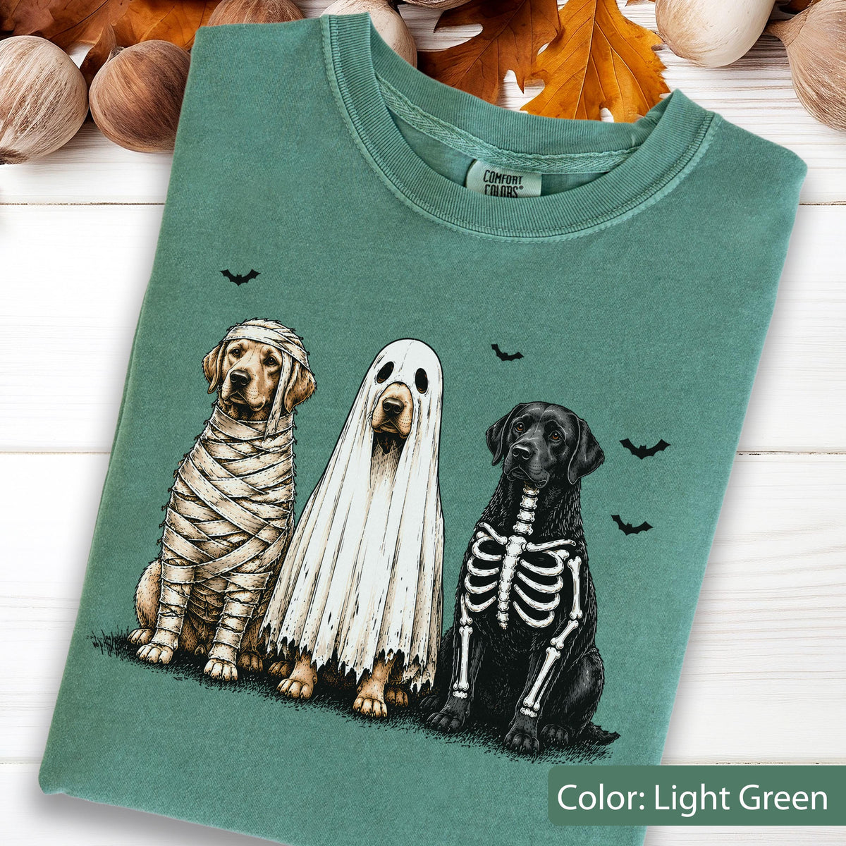 May include: A light green t-shirt featuring a graphic of three dogs in Halloween costumes: a golden retriever wrapped as a mummy, a golden retriever wearing a white ghost sheet, and a black labrador with a white skeleton print on its chest.