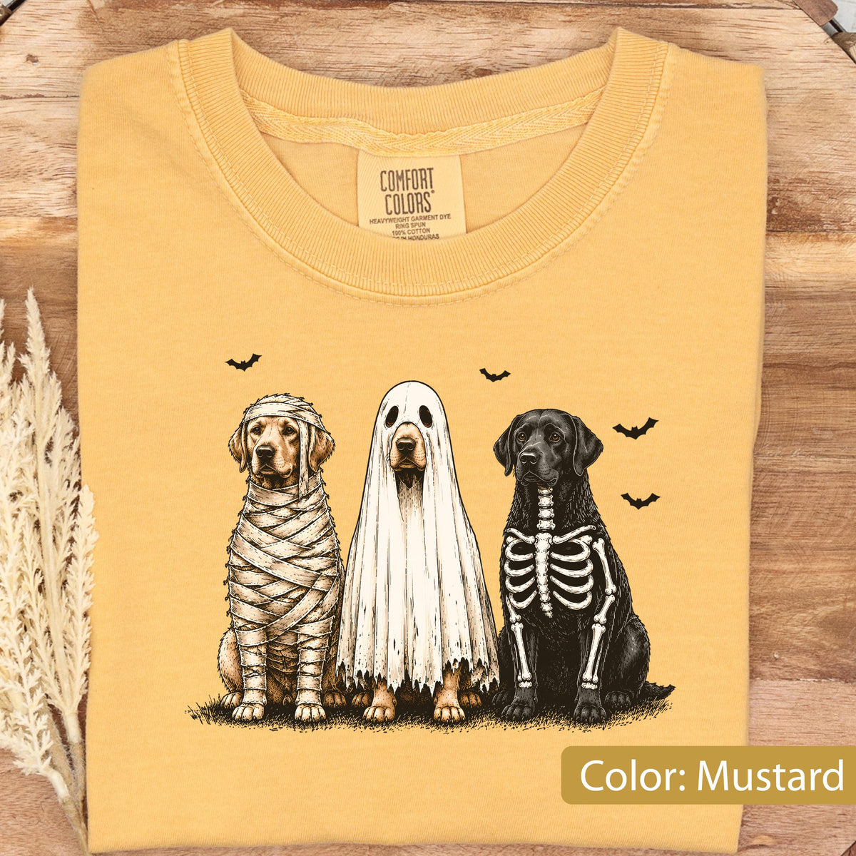 May include: A mustard yellow t-shirt featuring a graphic of three dogs in Halloween costumes: a golden retriever wrapped as a mummy, a dog wearing a white ghost sheet, and a black labrador with a white skeleton print. The Comfort Colors label is visible.