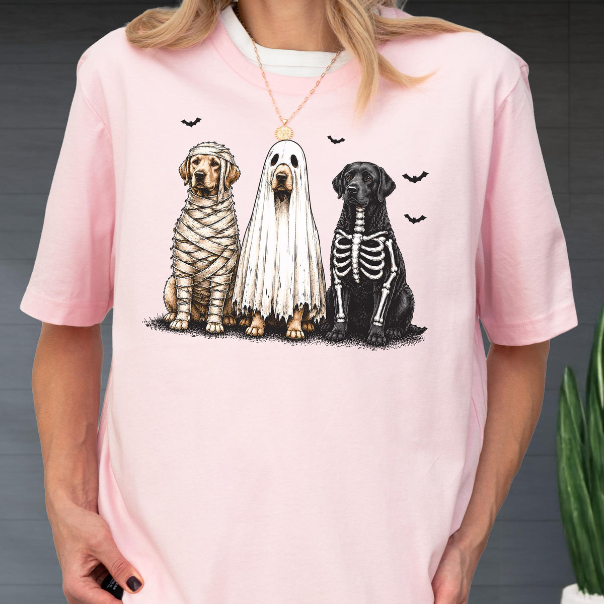 May include: A light pink t-shirt featuring a graphic of three dogs in Halloween costumes. One dog is wrapped in mummy bandages, one is wearing a white ghost sheet, and one has a skeleton print on its black fur, surrounded by small black bats.