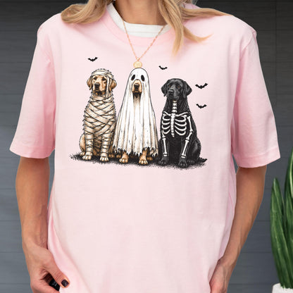 May include: A light pink t-shirt featuring a graphic of three dogs in Halloween costumes. One dog is wrapped in mummy bandages, one is wearing a white ghost sheet, and one has a skeleton print on its black fur, surrounded by small black bats.