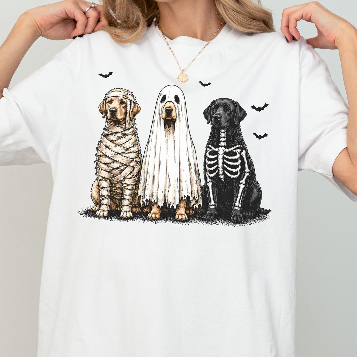 May include: A white t-shirt featuring a graphic of three dogs in Halloween costumes. One dog is wrapped in mummy bandages, one is wearing a white ghost sheet, and one has a skeleton print on its black fur. Small black bats fly above them.