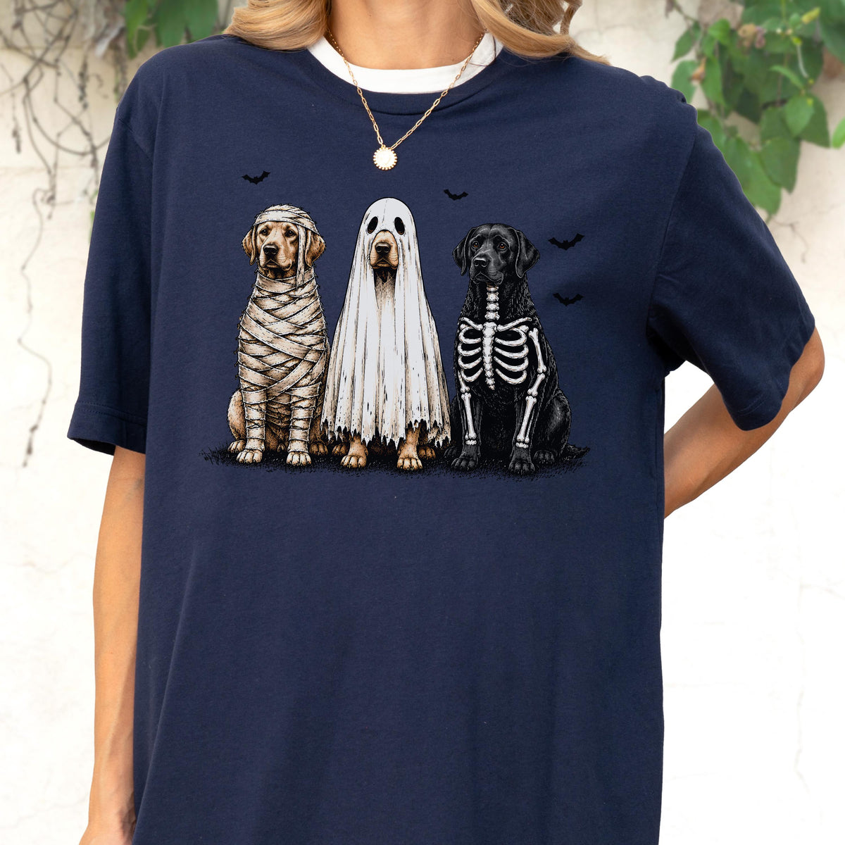 May include: A navy blue short-sleeve t-shirt featuring a graphic print of three dogs in Halloween costumes. One dog is wrapped like a mummy, one is wearing a white ghost sheet, and one has a skeleton ribcage design. Small black bats fly above.