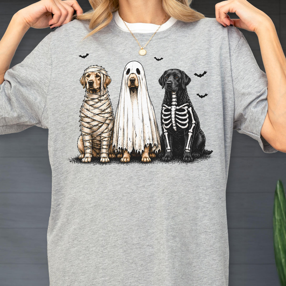May include: A heather gray t-shirt featuring a graphic of three dogs in Halloween costumes. One dog is wrapped in mummy bandages, one is wearing a white ghost sheet, and one has a white skeleton print on its black fur, with small black bats.