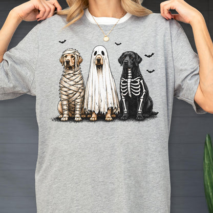 May include: A heather gray t-shirt featuring a graphic of three dogs in Halloween costumes. One dog is wrapped in mummy bandages, one is wearing a white ghost sheet, and one has a white skeleton print on its black fur, with small black bats.