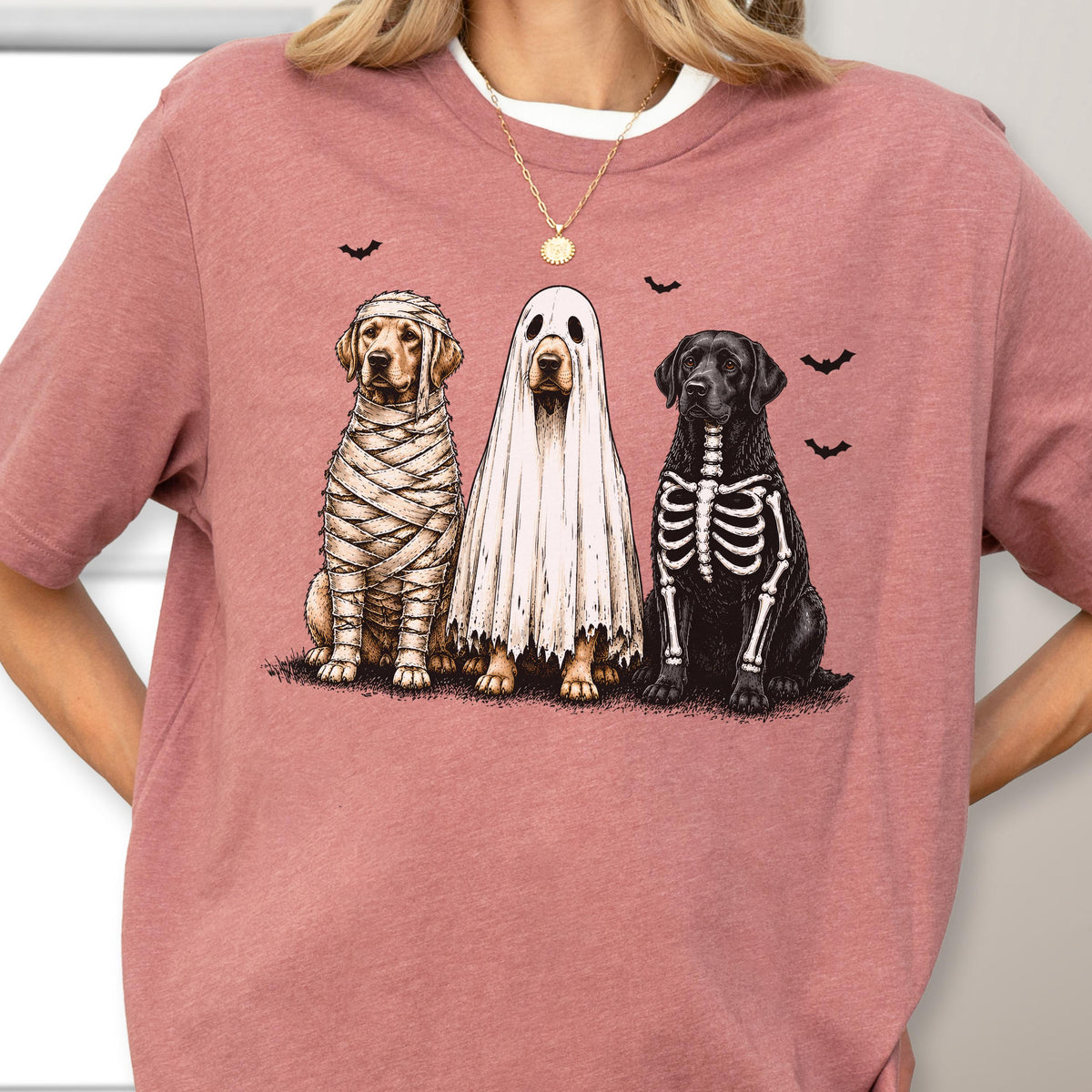 May include: A dusty rose short-sleeve t-shirt featuring a graphic of three dogs in Halloween costumes. One dog is wrapped in mummy bandages, one is wearing a white ghost sheet, and one has a skeleton print on its chest, with small black bats.