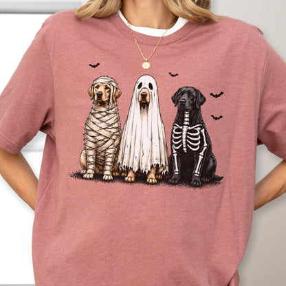 May include: A dusty rose short-sleeve t-shirt featuring a graphic of three dogs in Halloween costumes. One dog is wrapped in mummy bandages, one is wearing a white ghost sheet, and one has a skeleton print on its chest, with small black bats.