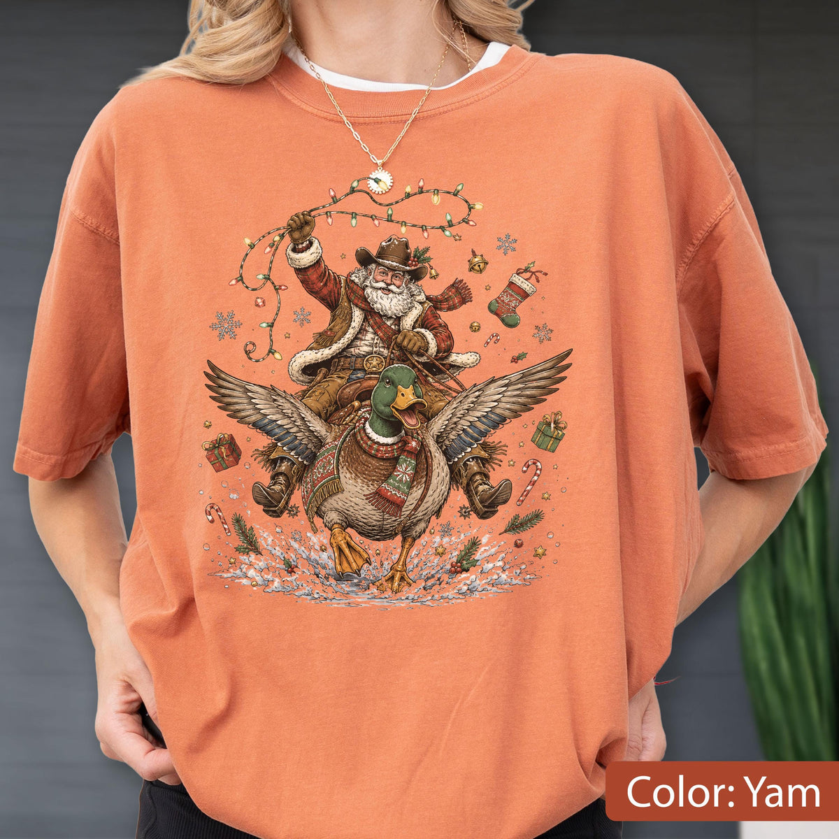 May include: A burnt orange short-sleeve t-shirt featuring a graphic print of Santa Claus wearing a cowboy hat and riding a flying mallard duck. The design includes Christmas lights, snowflakes, gift boxes, and candy canes surrounding the figures.