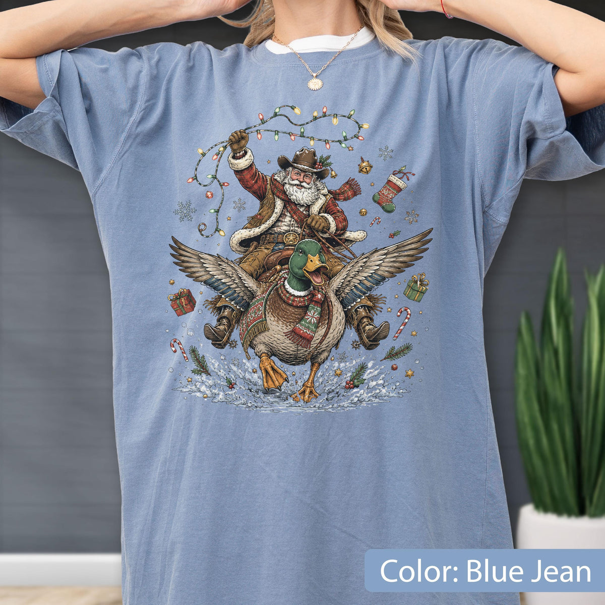 May include: A blue jean colored t-shirt featuring a graphic illustration of Santa Claus wearing a cowboy hat and scarf while riding a flying mallard duck. Santa holds a string of colorful Christmas lights, surrounded by snowflakes, gifts, and candy canes.